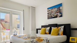 Ein Schlafzimmer mit einem Bett, einem Nachttisch, einem Gemälde und Blick auf Gebäude durch ein Fenster.