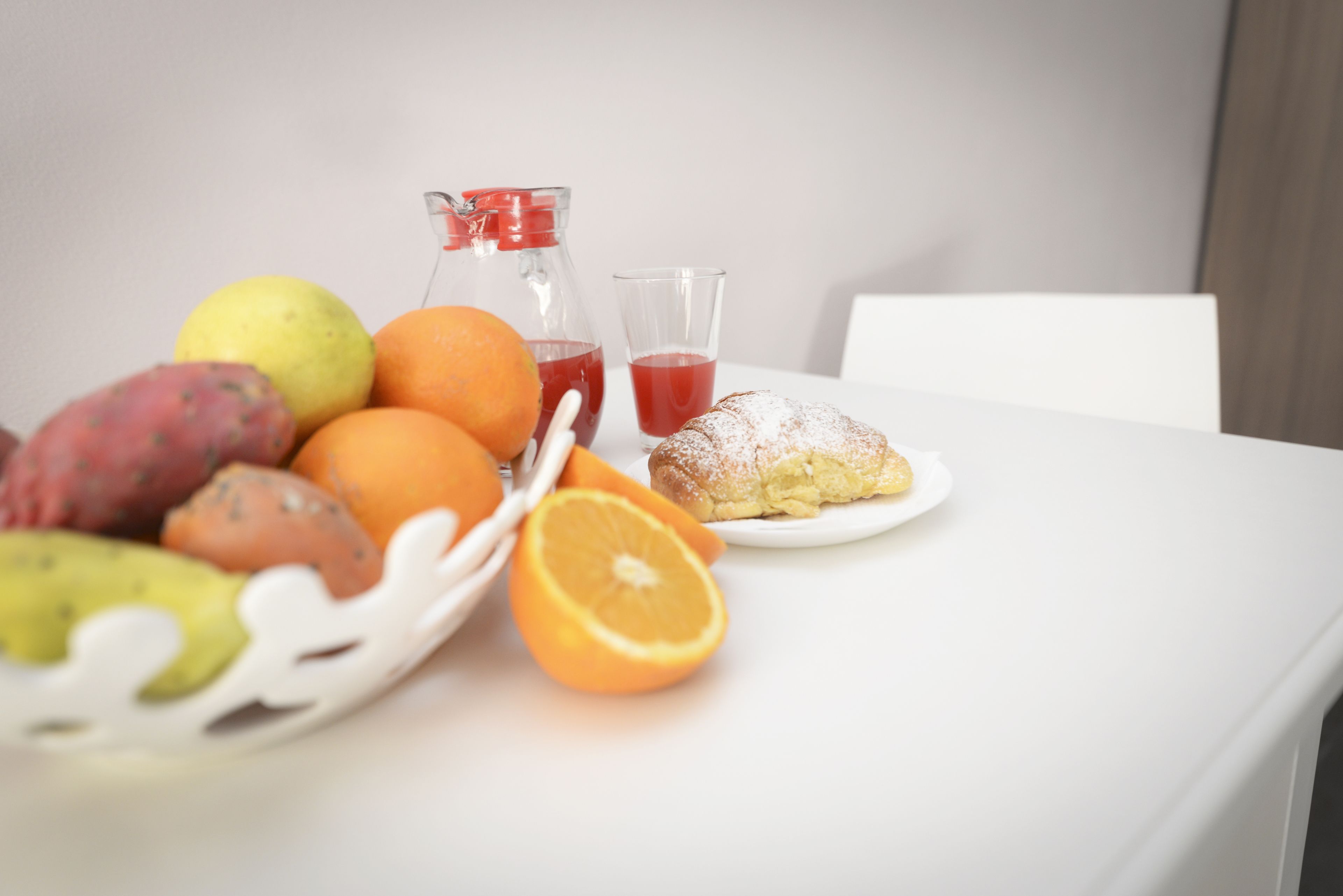 A table with a fruit basket containing oranges, a lemon, and a kiwi, a glass of red juice, a glass of water, and a pastry.