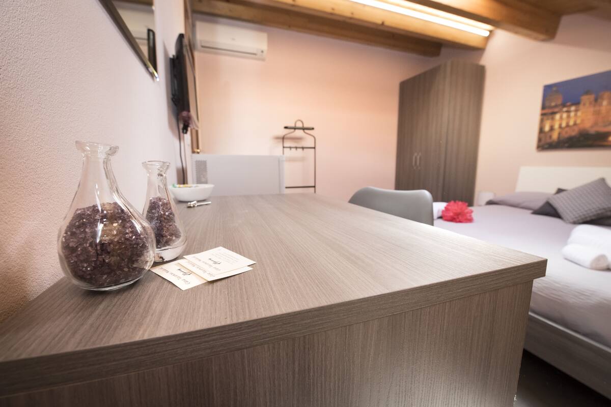 Een hotelkamer met een houten receptiebalie, twee glazen vazen met gedroogde bloemen, een wit wandgemonteerde airconditioner, een bed met een grijs hoofdeinde en een schilderij aan de muur.
