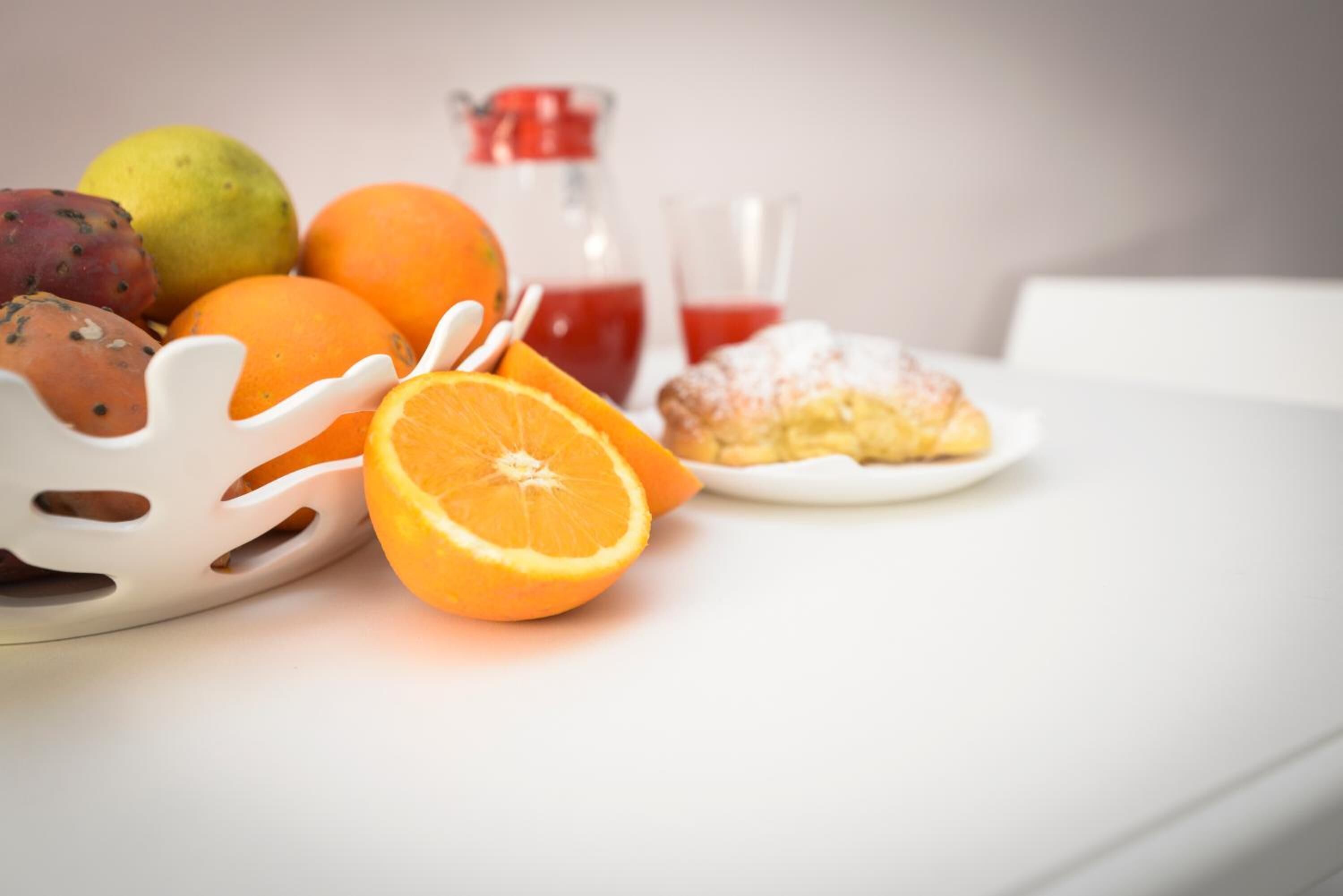 A fruit bowl with oranges and a pear, a glass of red juice, and a pastry on a white table.