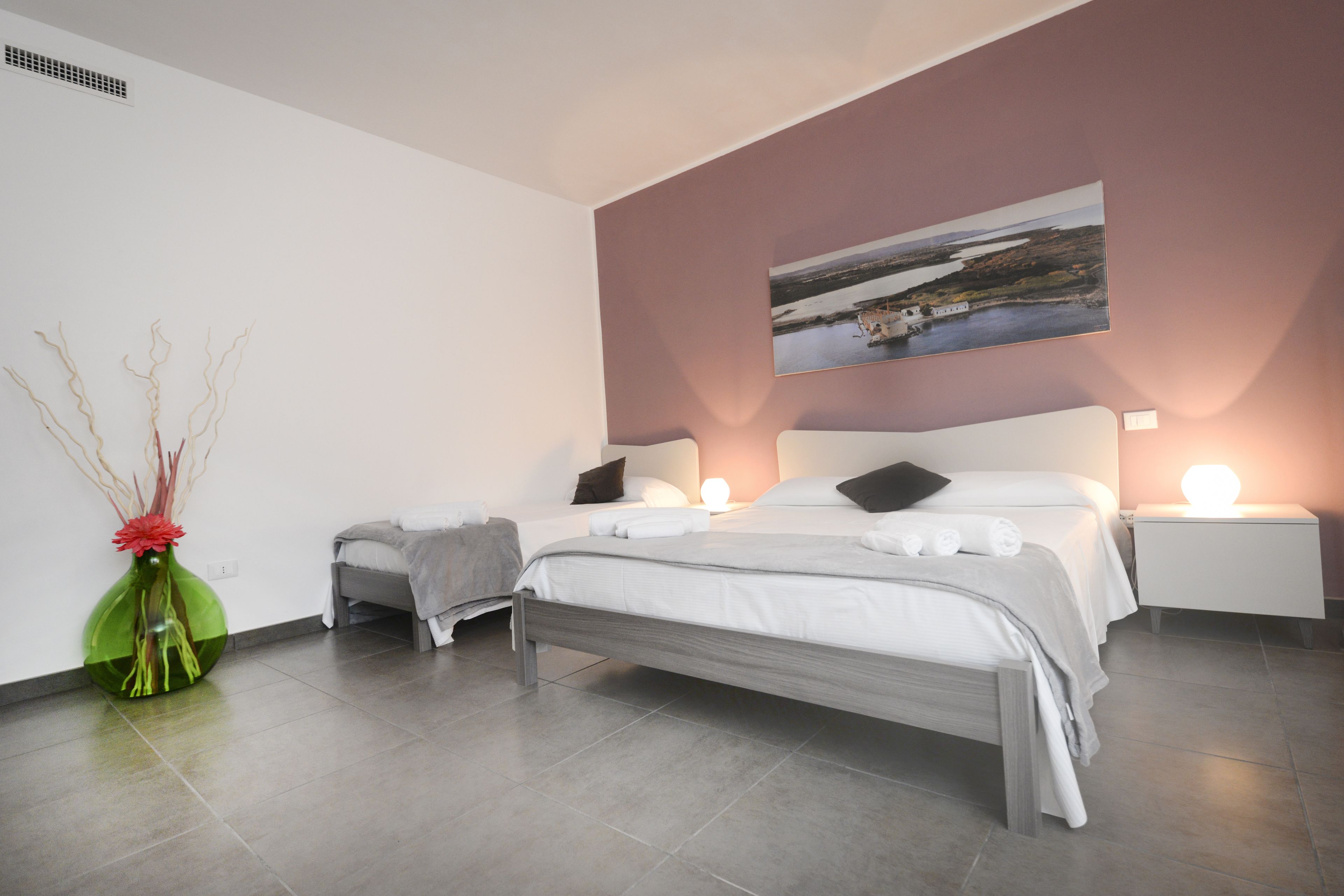 A modern bedroom with a large bed, bedside tables, a nightstand, a wall-mounted painting, and a decorative vase.