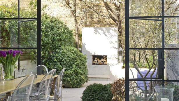 Un coin repas avec une table et des chaises, un vase de fleurs et une vue sur un jardin, visible à travers de grandes fenêtres.