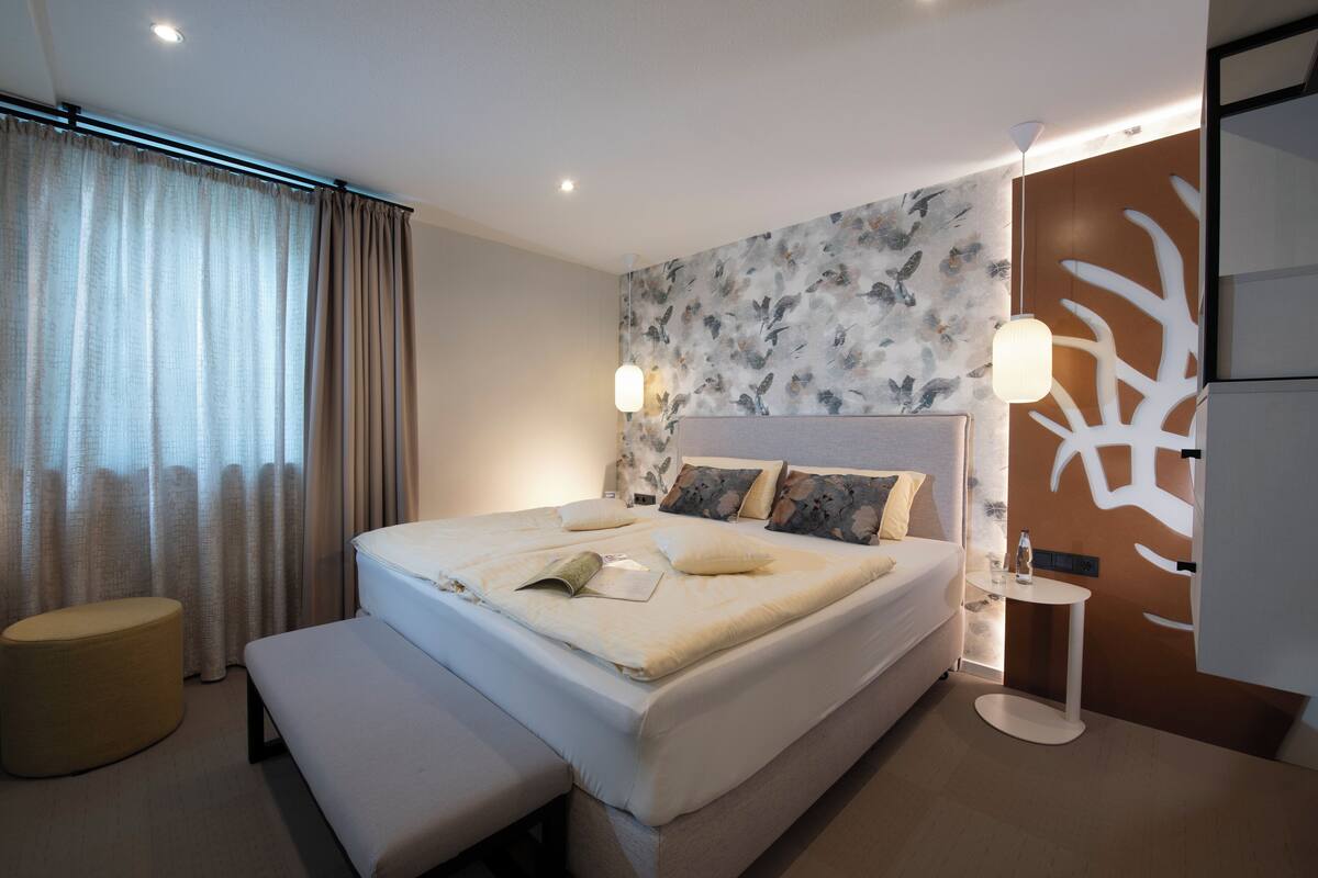 Quarto moderno com uma cama grande, um banco, uma mesinha e um painel decorativo.