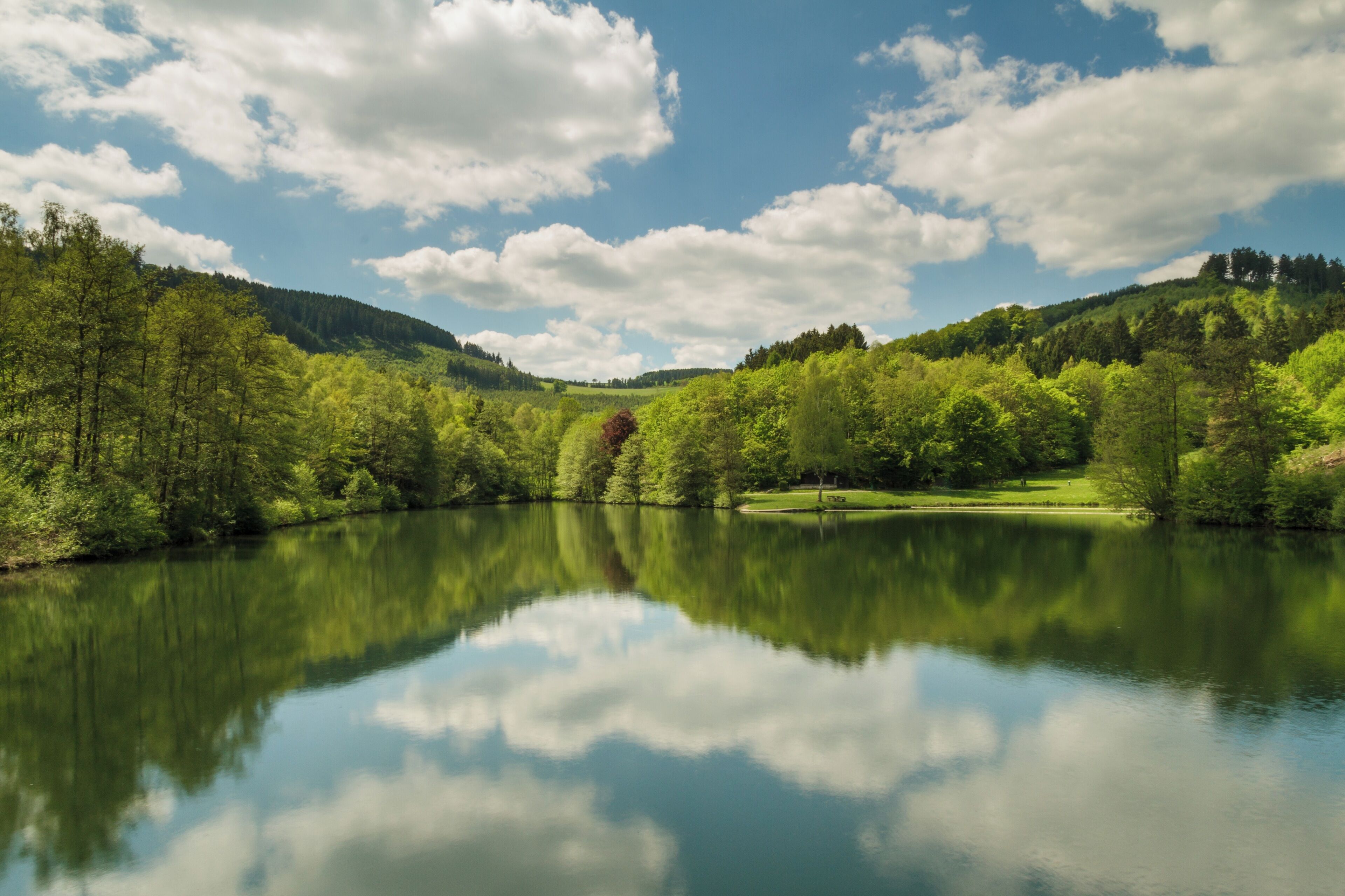 Um lago tranquilo rodeado por vegetação exuberante e árvores, refletindo o céu e as nuvens.