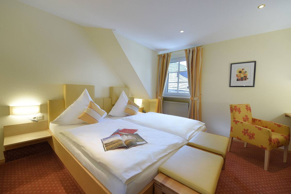 Quarto de hotel com uma cama grande, mesas de cabeceira, uma poltrona e uma janela com cortinas.