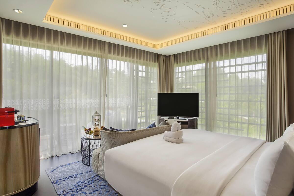 Une chambre d’hôtel moderne dotée d’un grand lit, d’un téléviseur à écran plat, d’une table d’appoint ronde avec une lampe, et offrant une vue sur la verdure par les fenêtres.