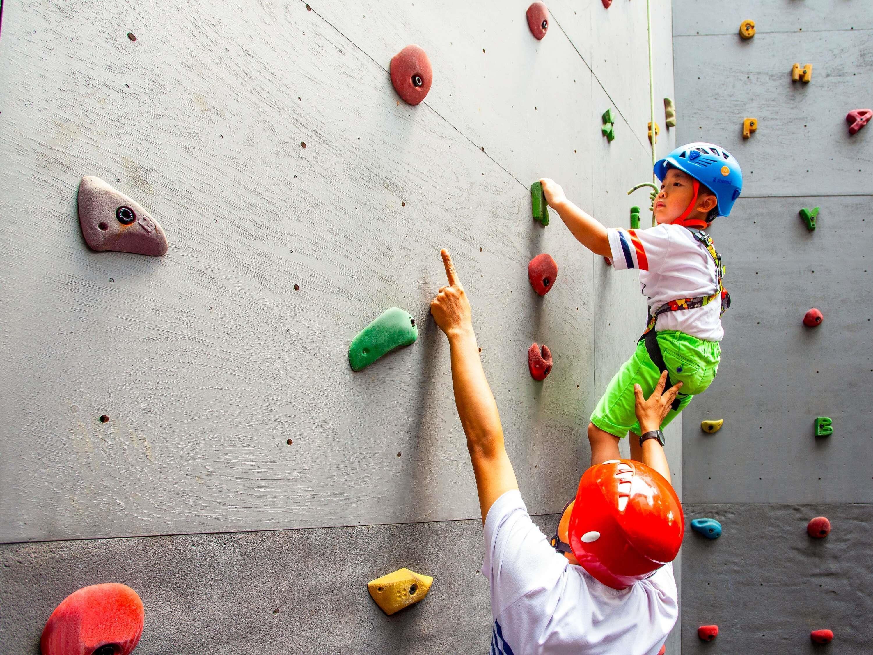 Criança escalando uma parede de escalada com pontos de apoio de cores variadas, com a supervisão de um adulto.