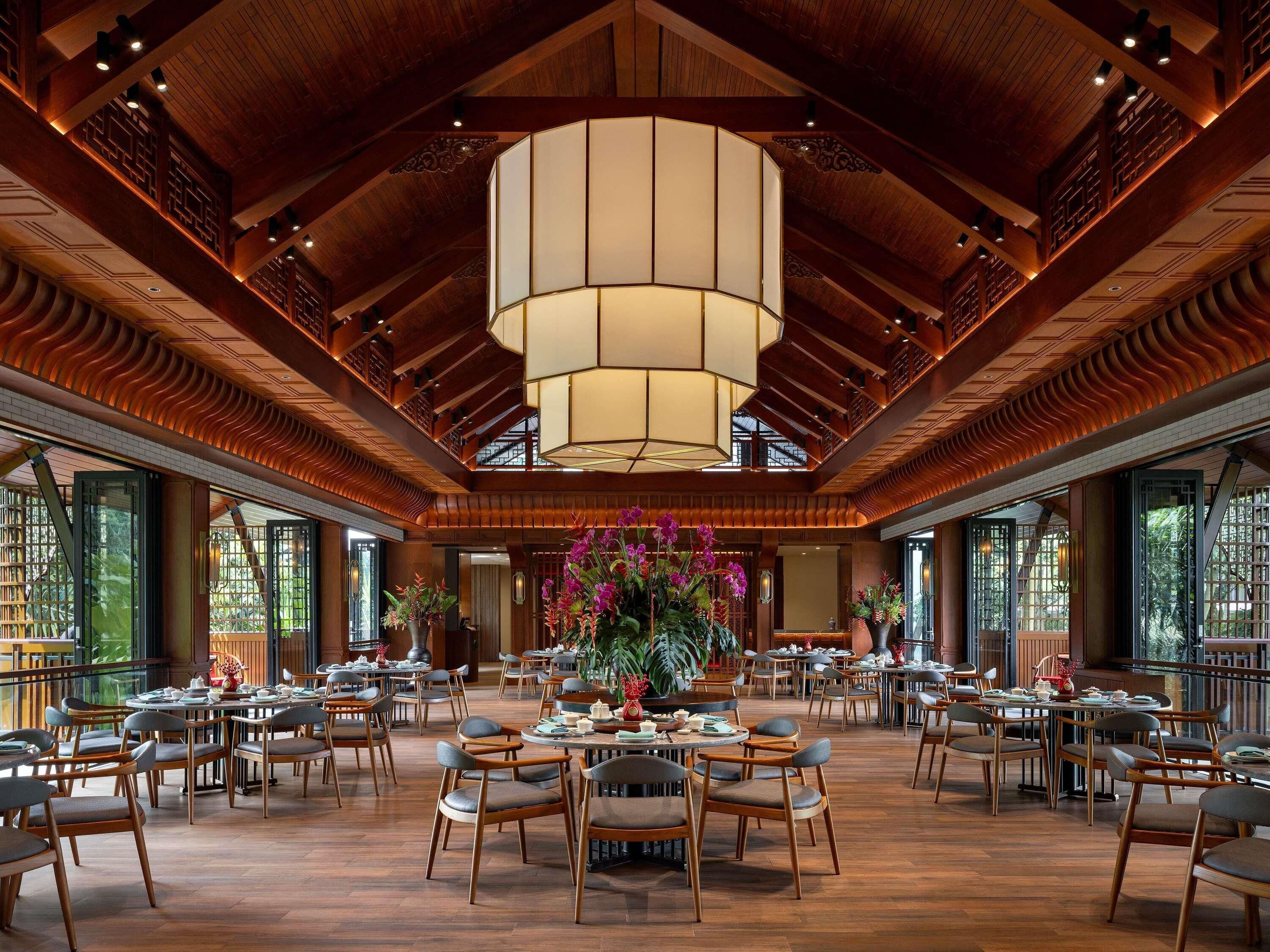 A spacious dining area with wooden tables and chairs, a large central chandelier, and large windows on the sides.