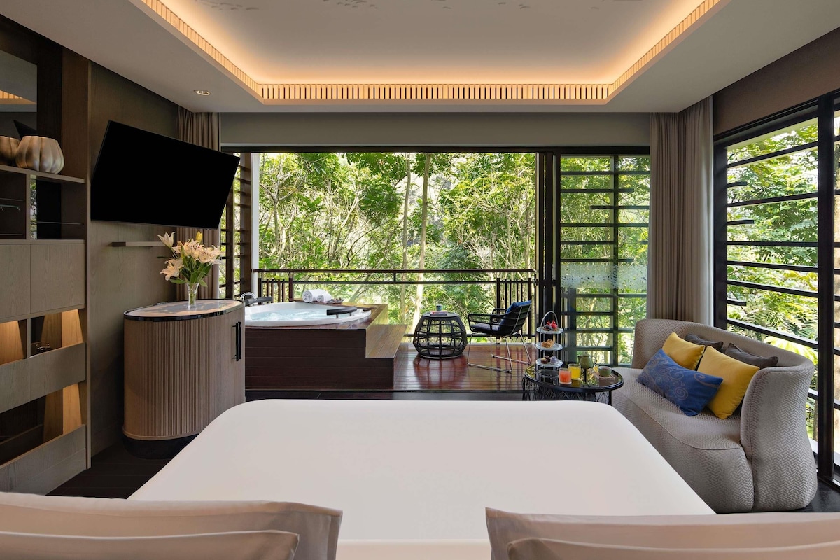 Una habitación de hotel moderna con una cama grande, un área de estar con sofá y sillones, una pequeña mesa con hidromasaje y un televisor montado en la pared.