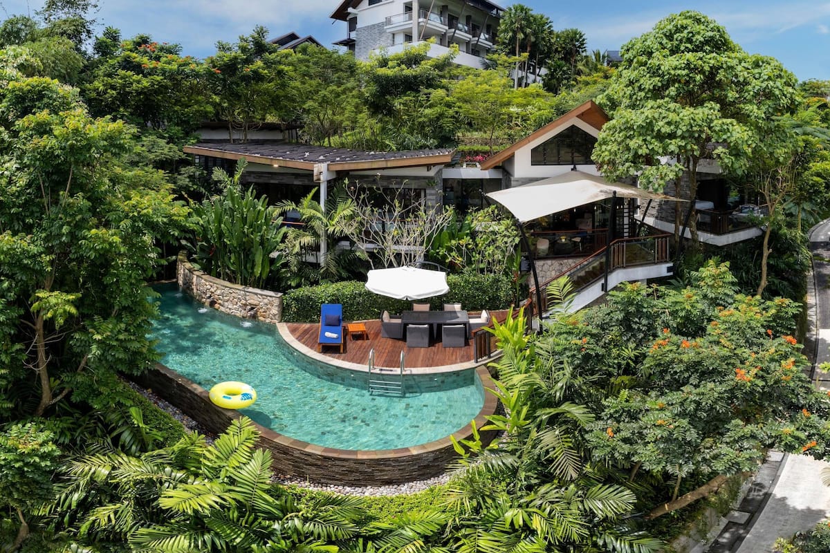 Un complexe hôtelier moderne doté d’une piscine circulaire entourée d’une végétation luxuriante, d’un espace extérieur aménagé et couvert, et d’un bâtiment à plusieurs étages avec des balcons.