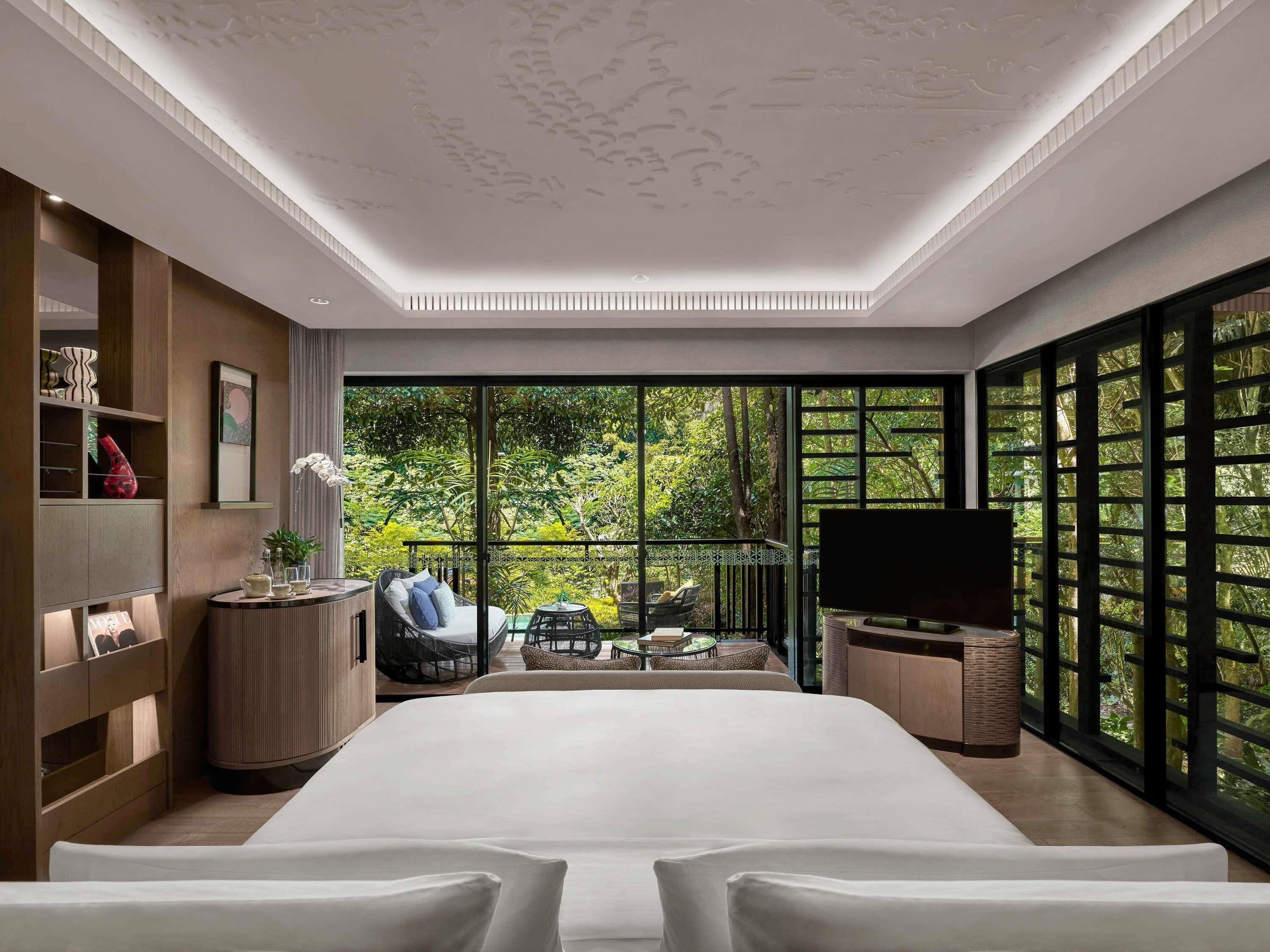 A modern hotel room with a large bed, a TV, a balcony with a view of greenery, and a wooden cabinet with decorative items.