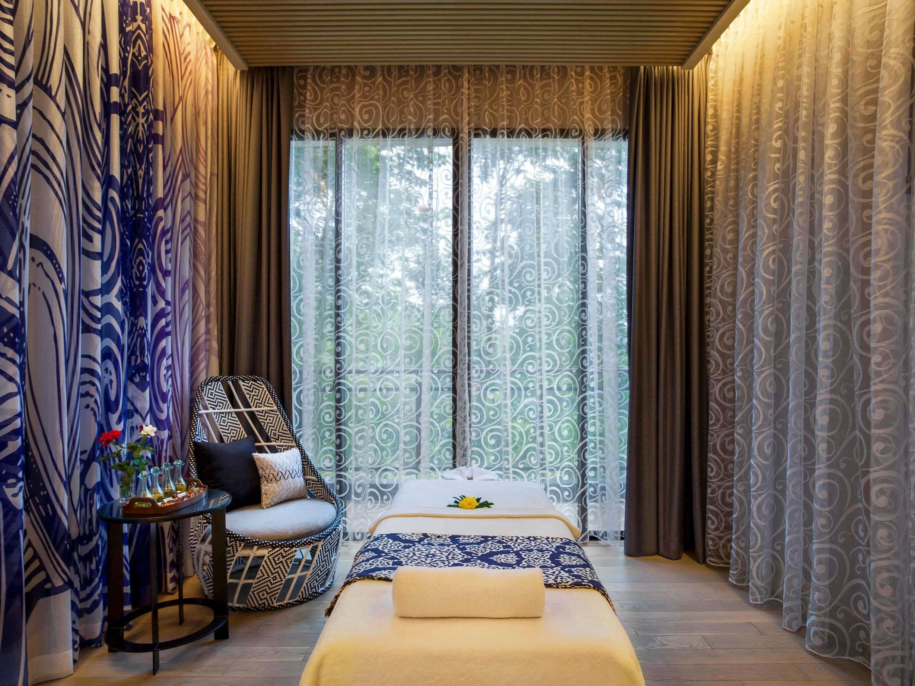 A bedroom with a bed, a small table with decorative items, a chair, and a window with curtains.