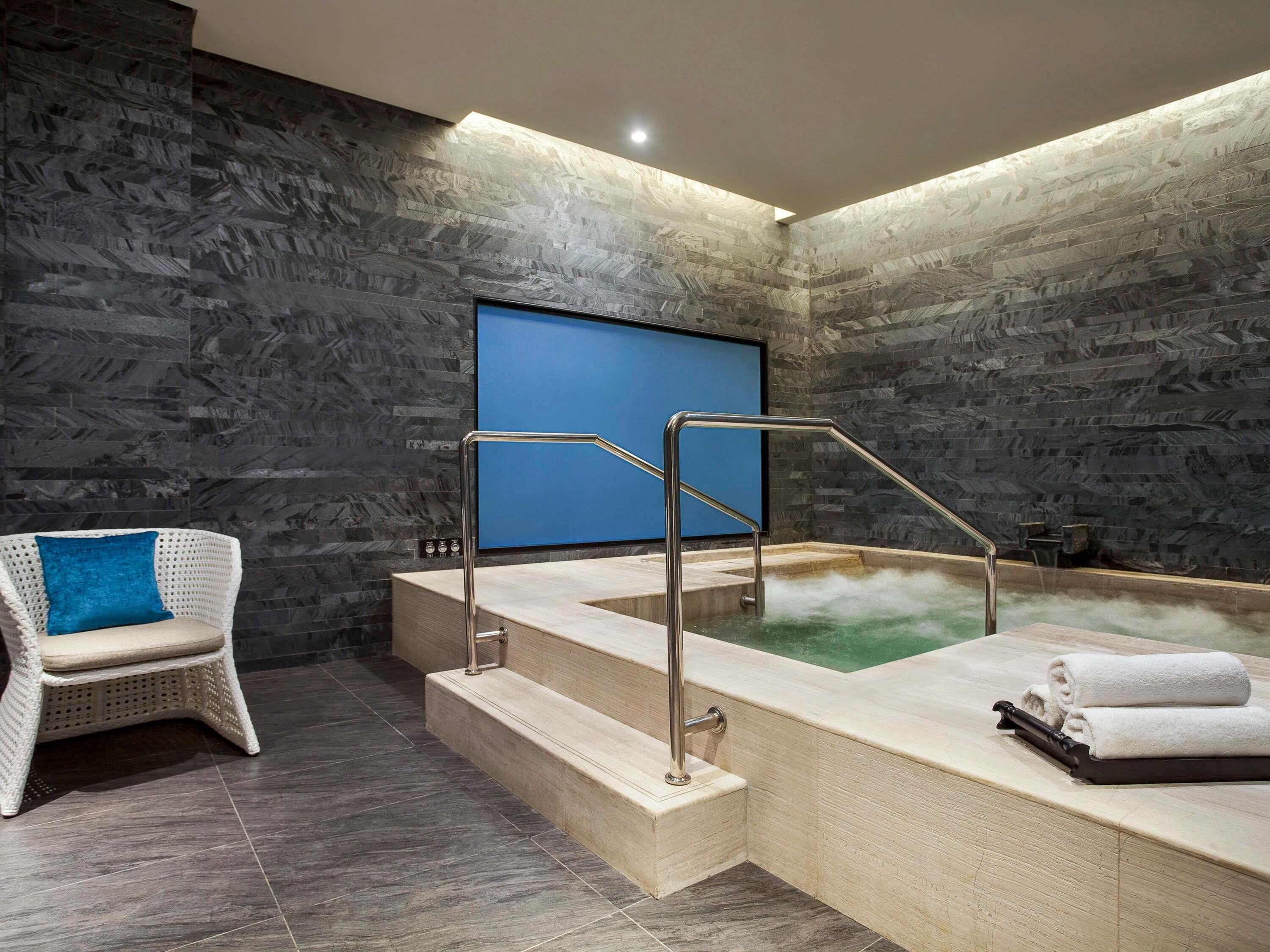 A modern spa room with a stone wall, a blue screen, a white wicker chair, and a hot tub.