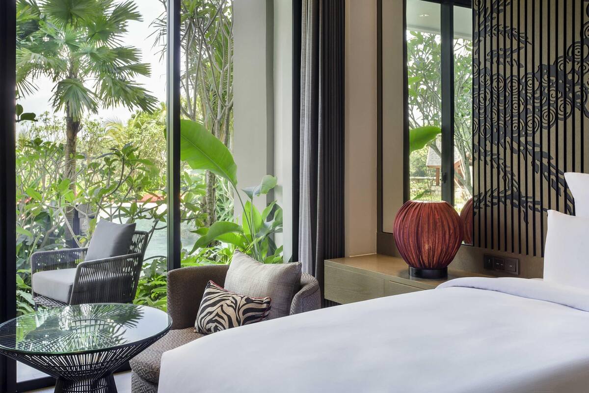 Une chambre d’hôtel moderne dotée d’une grande fenêtre, d’un lit, d’une table ronde en verre et d’un coin salon avec des fauteuils.