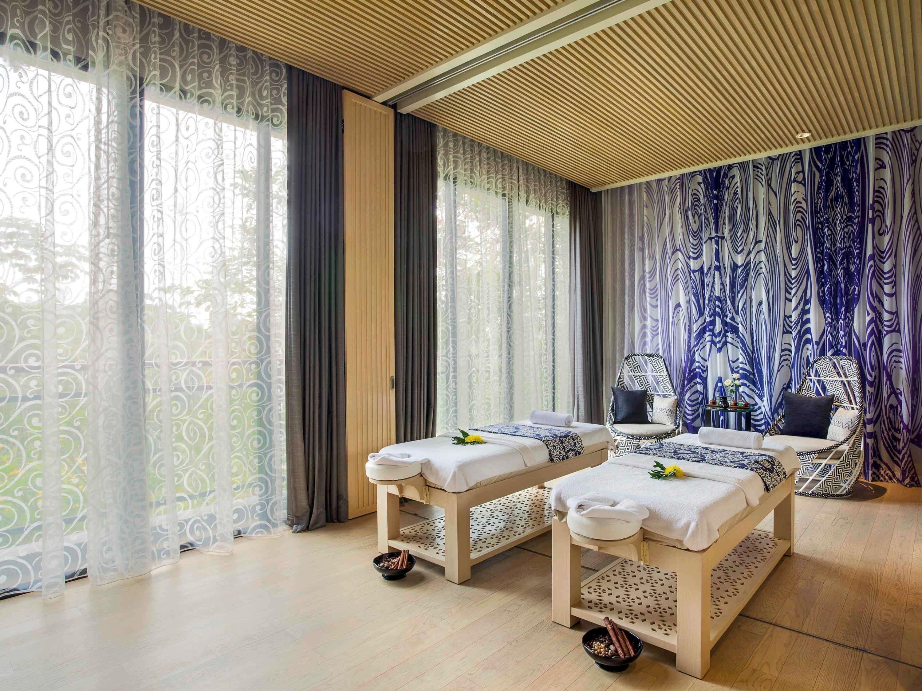 Een sparuimte met twee massagetafels, een stoel en een groot raam met decoratieve gordijnen.