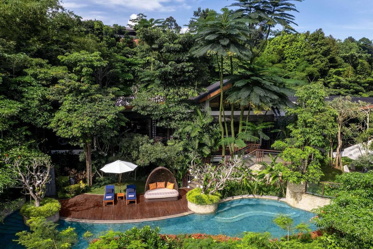 Un complexe hôtelier luxuriant doté d’une piscine entourée d’une végétation tropicale dense, d’une terrasse en bois avec des chaises longues et, au fond, d’un bâtiment de style traditionnel.