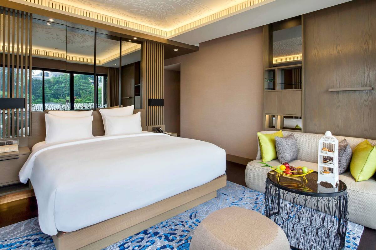 Une chambre d’hôtel moderne, dotée d’un grand lit, d’un canapé, d’une table basse ronde et offrant une vue sur la verdure environnante.