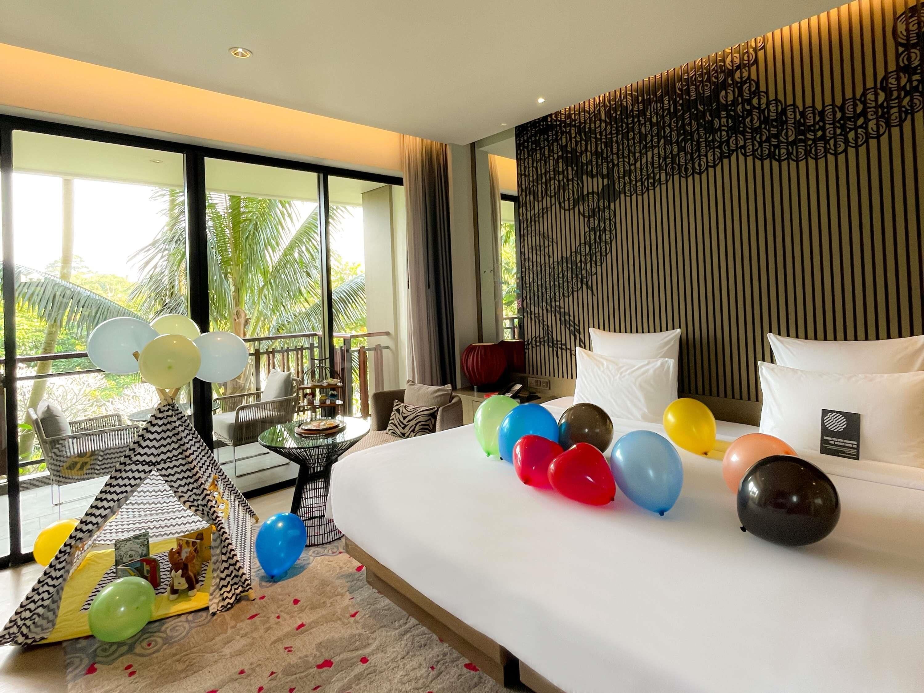 Ein Hotelzimmer mit Bett, bunten Luftballons, einer kleinen Spielecke und einem Balkon mit Blick auf Palmen.
