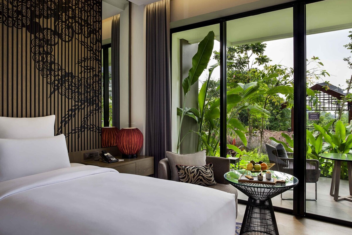 Een moderne hotelkamer met een groot bed, een nachtkastje met telefoon, een glazen salontafel en uitzicht op de tuin door grote ramen.