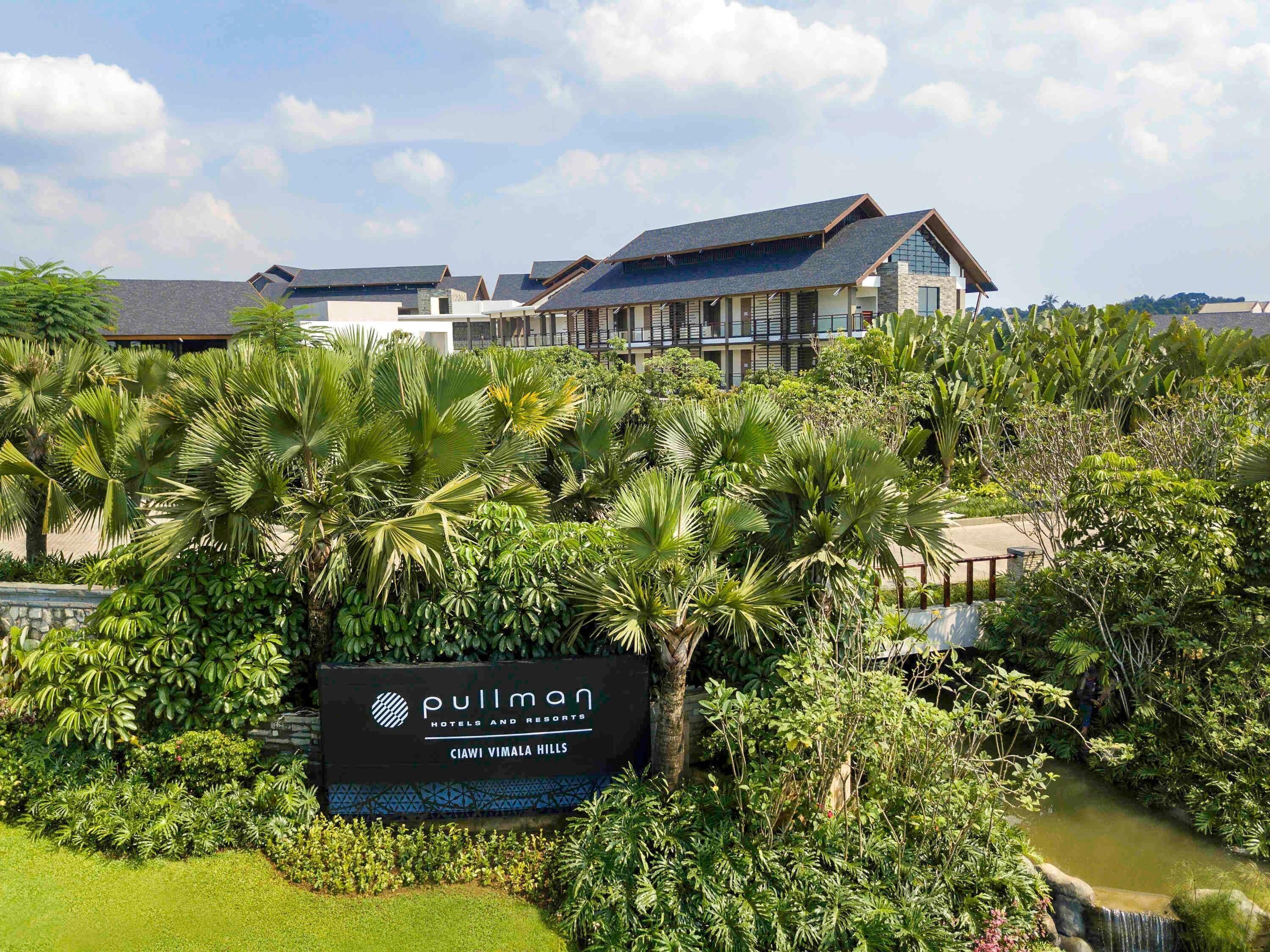 Señal de hotel con la inscripción "Pullman Hotels and Resorts" y "Ciawi Vimala Hills", rodeada de exuberante vegetación y una pequeña cascada.