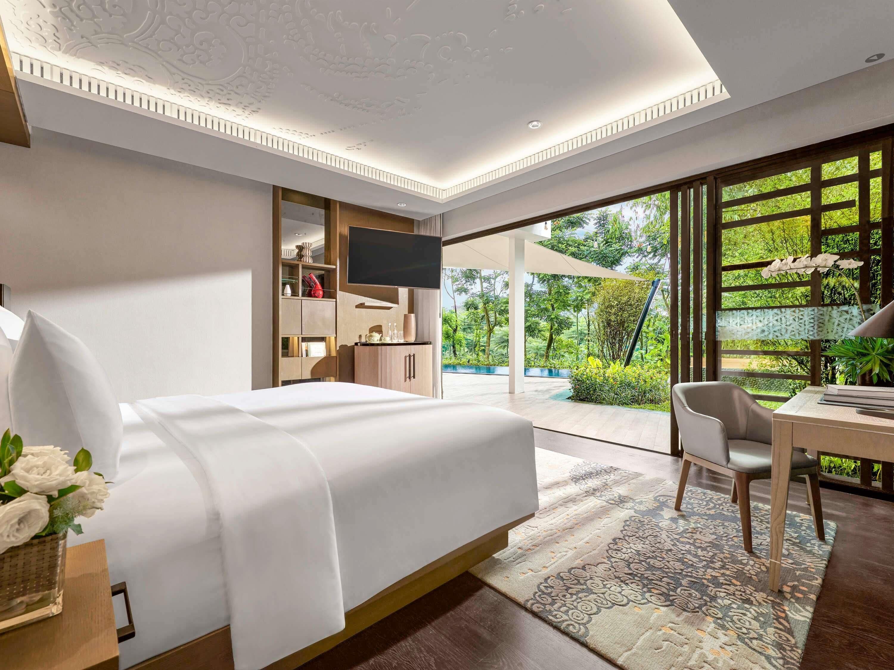 A modern hotel room with a bed, a desk, a chair, a TV, and a view of greenery outside.