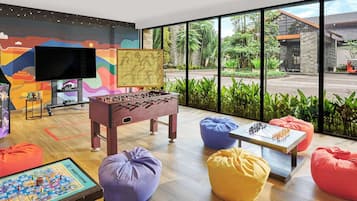 Relax in a spacious room with a large window offering a view of the garden and a building outside, featuring a flat-screen TV, foosball table, and colorful mural.