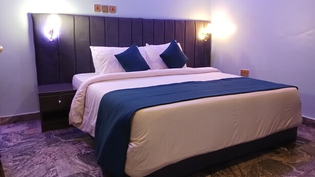 A neatly made bed with a dark headboard, a bedside table, and two wall-mounted light fixtures. - Degok Hotel (Abuja)