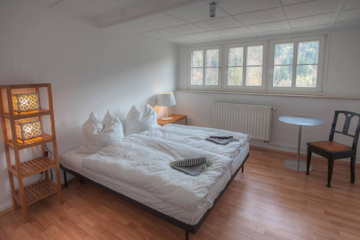 A bedroom with a bed, a bedside table with a lamp, a chair, a small table, and a window with a view of trees.