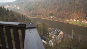 A view of a river with a wooden railing in the foreground, a building with a sloped roof, and a forested hillside in the background. - Die Burg Schöna - In a national park (Reinhardtsdorf-Schoena)