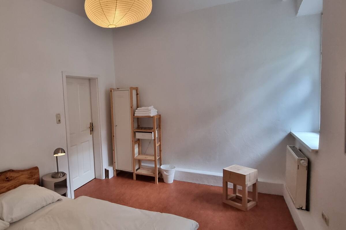 A bedroom with a wooden headboard bed, a bedside lamp, a wooden ladder shelf, a radiator, and a window with a view.