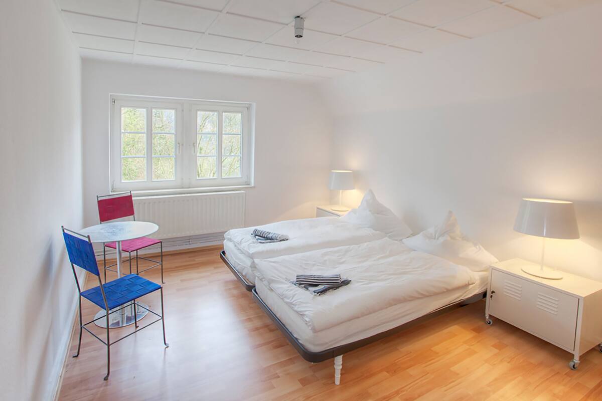 A room with two beds, a small table, and a chair. There is a window with blinds, a radiator, and two bedside lamps.