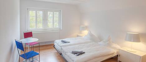 Quarto com duas camas, uma mesinha e uma cadeira. Há uma janela com persianas, um radiador e duas luminárias de cabeceira.