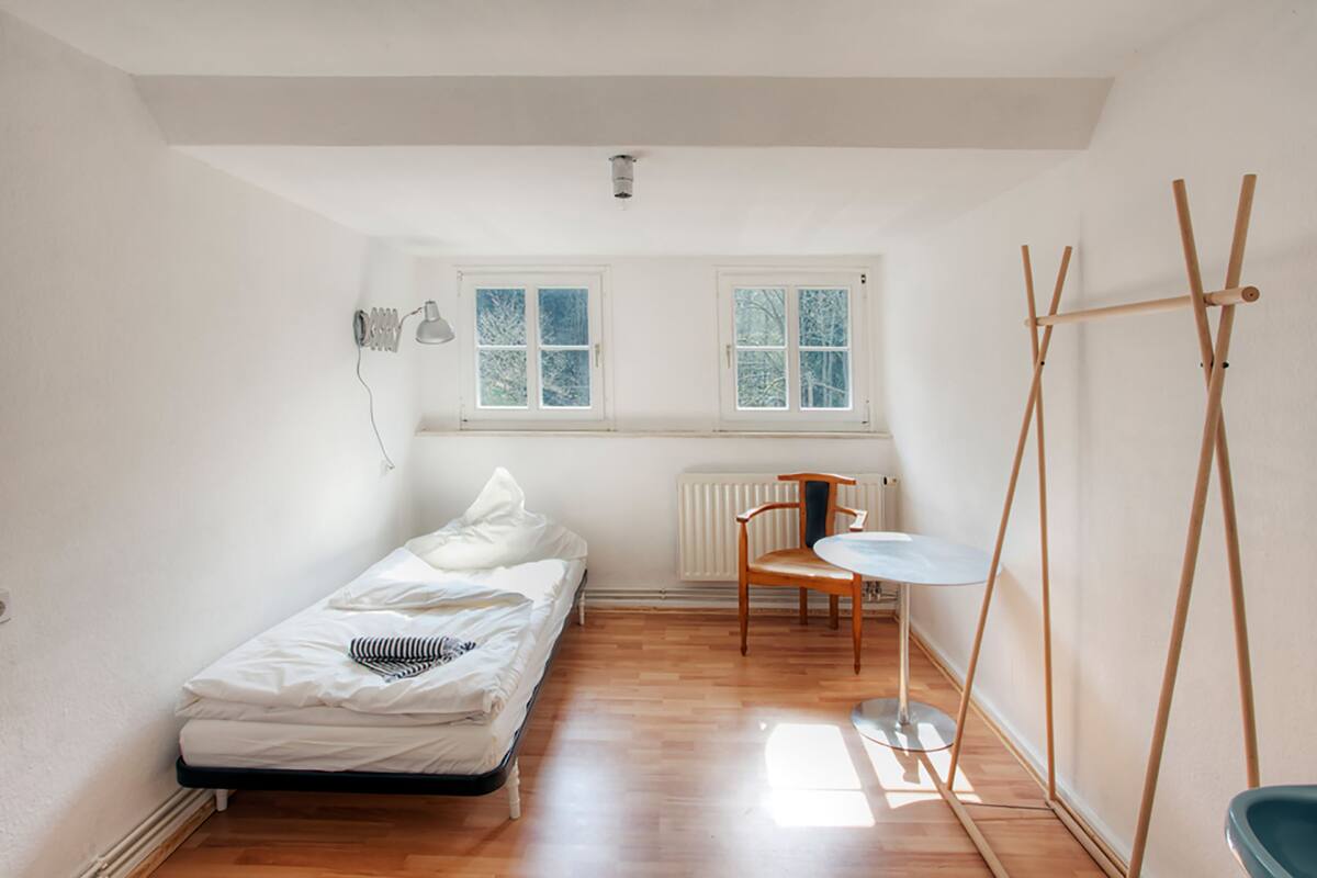 A small, bright room with a single bed, a chair, a table, and a wooden stand.