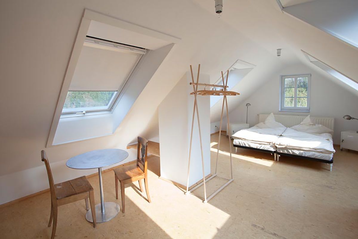 Et rum med højt til loftet, ovenlys, en seng, et bord med stole og en hængende lampe.