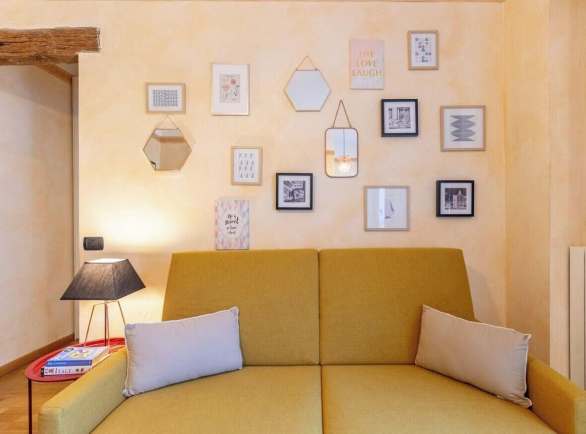 Een kamer met een geel sofa, een klein bijzettafeltje met een lamp, en een wand versierd met diverse ingekaderde foto's en een zeshoekige spiegel.