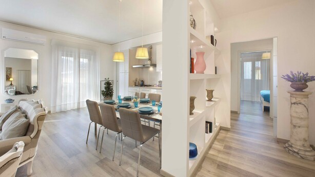 Un espace de salle à manger moderne, avec une table dressée pour quatre personnes, entourée de chaises. Un mur blanc avec des étagères se trouve à droite, et un couloir mène à une autre pièce.