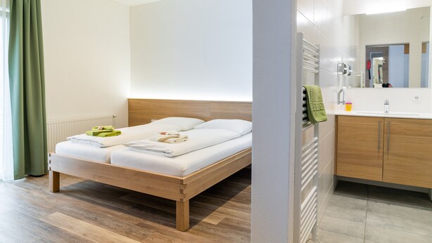 Una habitación de hotel moderna con una cama de madera, un baño con espejo y una cortina verde.