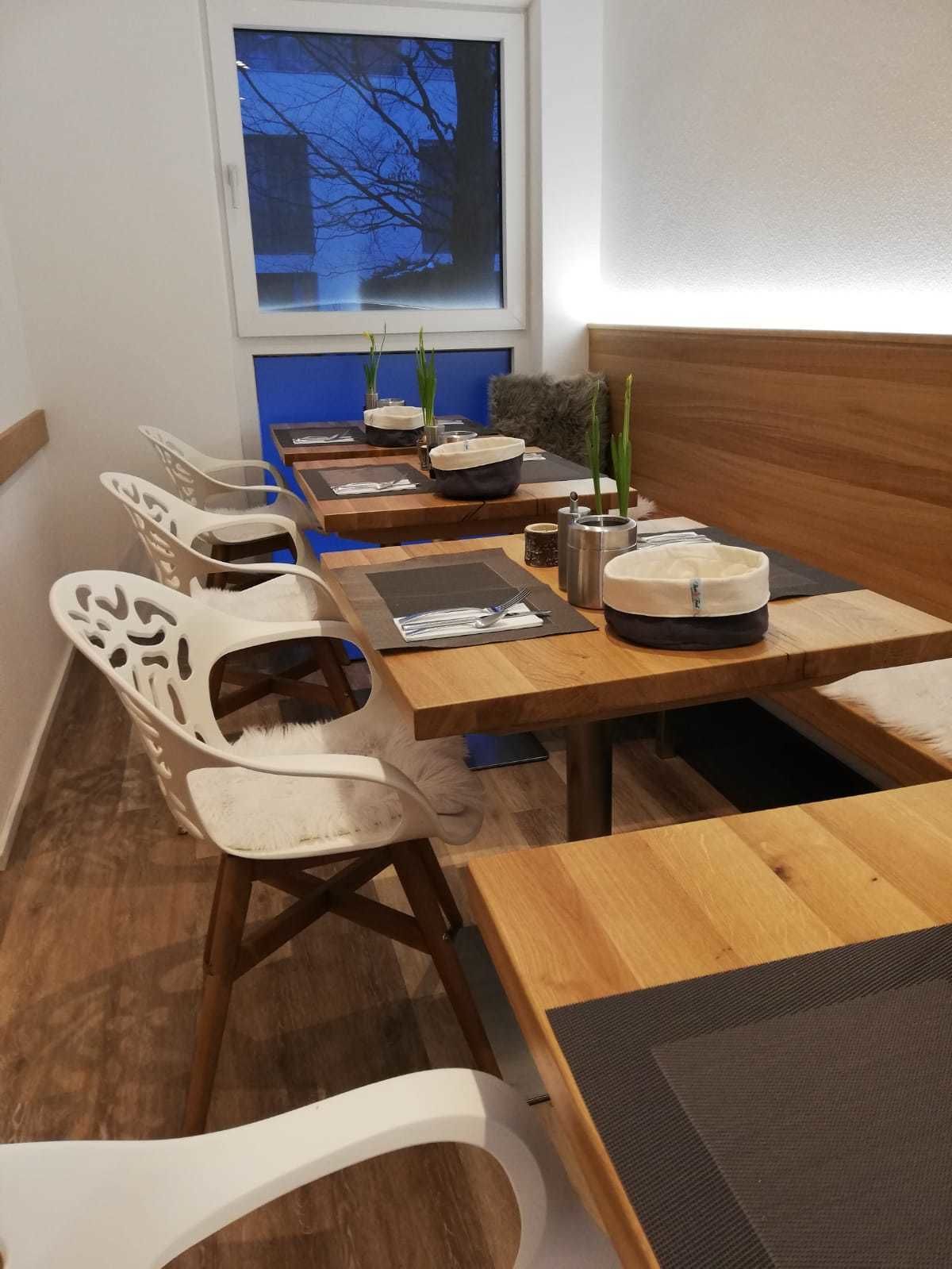 Un comedor con mesas de madera y sillas blancas, algunas con diseños recortados. Hay manteles individuales negros y un camino de mesa blanco. Se ve una ventana con vista a árboles desnudos.