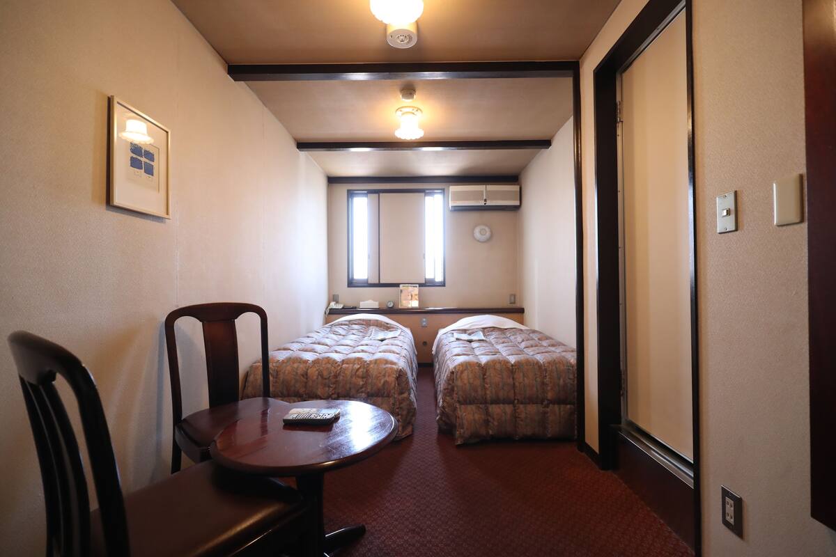 A hotel room with two beds, a small table, a chair, a window, and a ceiling light.