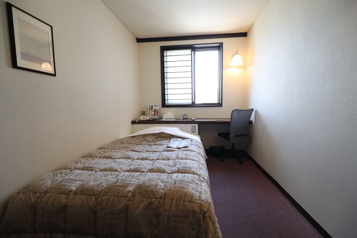 A small, single-bed room with a desk and chair, a window with blinds, and a wall-mounted lamp.