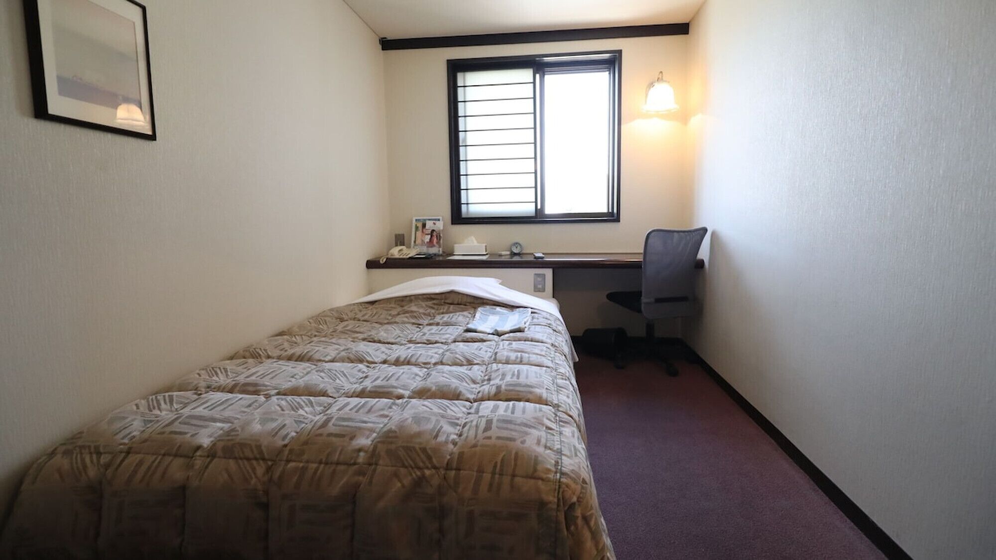 A bedroom with a bed, a desk, a chair, a window, and a wall-mounted light fixture.