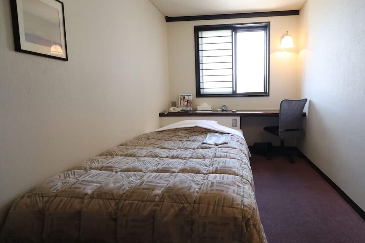 A bedroom with a bed, a desk, a chair, a window, and a wall-mounted light fixture.