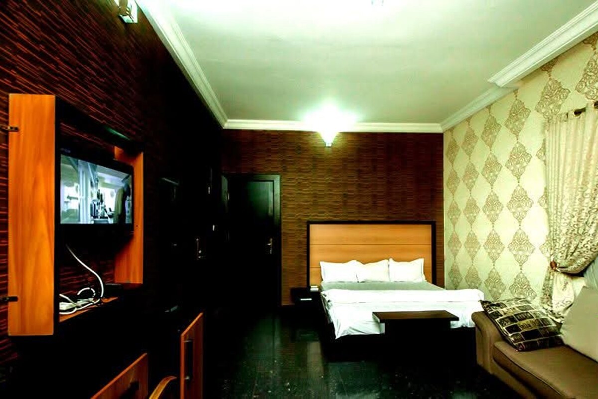 A hotel room with a wooden wall, a bed with white linens, a television, and a sofa with patterned cushions.