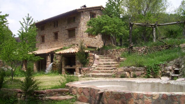Un bâtiment rustique en pierre, avec un toit en tuiles, un escalier en pierre menant à l’entrée et une petite fontaine en premier plan.