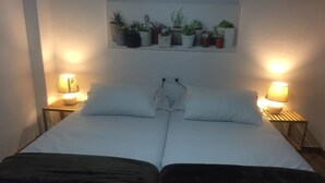 Un lit double avec du linge de lit blanc, deux tables de chevet avec des lampes et une étagère à plantes fixée au mur, située au-dessus.