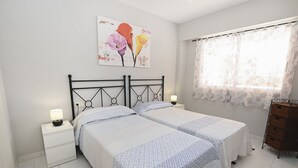 Ein Schlafzimmer mit zwei Betten, einem Nachttisch mit Lampe, einem Wandbild und einem Fenster mit Vorhängen.