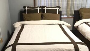 Un lit double avec une couvre-lit blanche et marron, deux oreillers et, au fond, une fenêtre avec des stores.