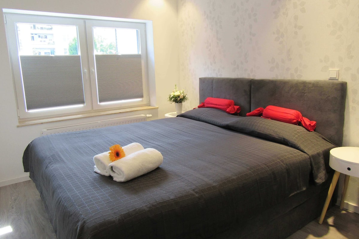 Ein Schlafzimmer mit einem großen Bett, einem Fenster mit Jalousien, einem Nachttisch mit Pflanze und einem Beistelltisch mit Lampe.