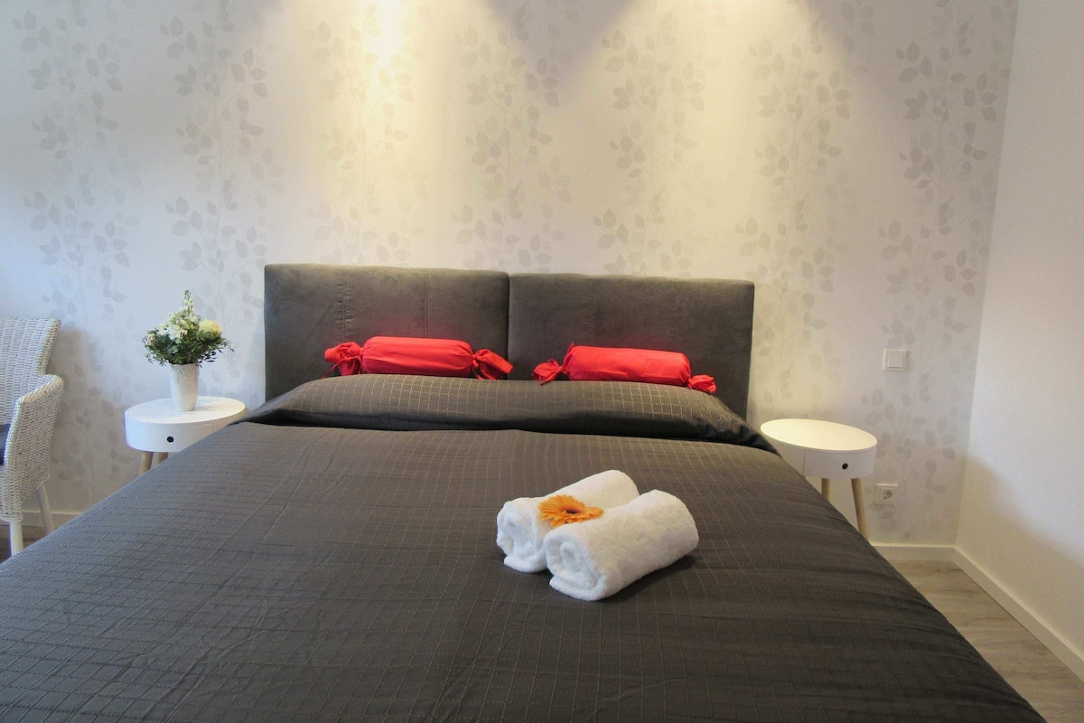 Ein ordentlich bezogenes Bett mit dunkler Tagesdecke und zwei roten Kissen, ein kleiner weißer Nachttisch mit Blumenvase sowie eine Wand mit dezenter Blattmusterung.