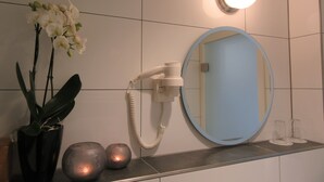 A bathroom with a white tiled wall, a round mirror, a hairdryer, and decorative items including a plant and two spherical objects. - Minsu Hotel (Muelheim an der Ruhr)
